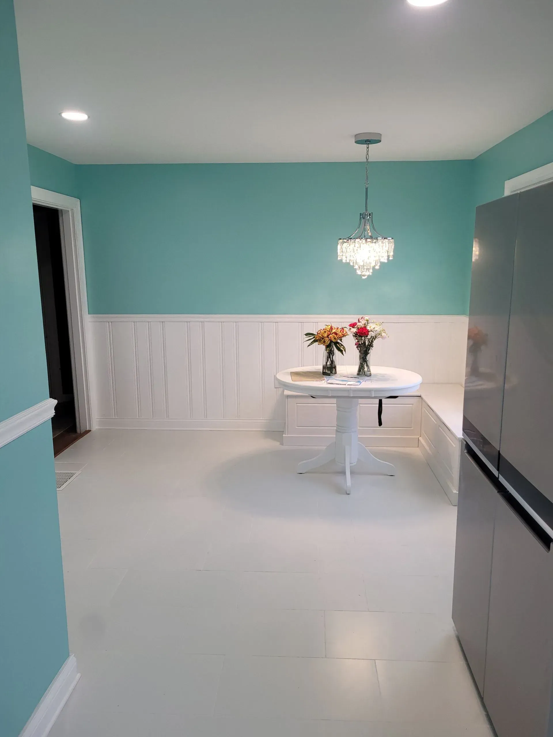 A bright, teal-painted dining area with white floors, wainscoting, and a round table topped with flowers.