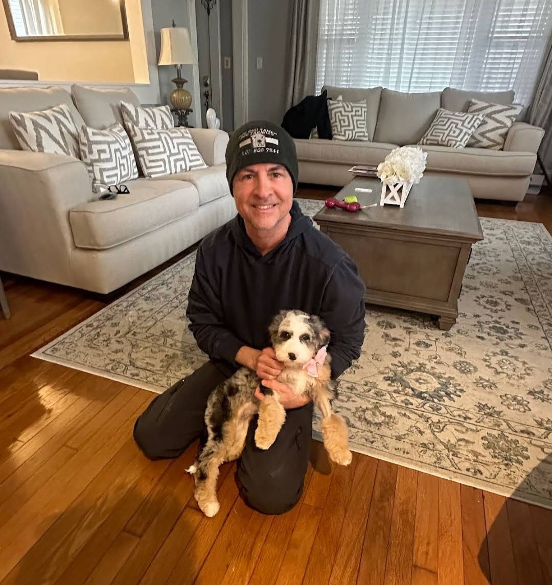 Person kneeling on hardwood floor, smiling and holding a small dog in a bright living room.