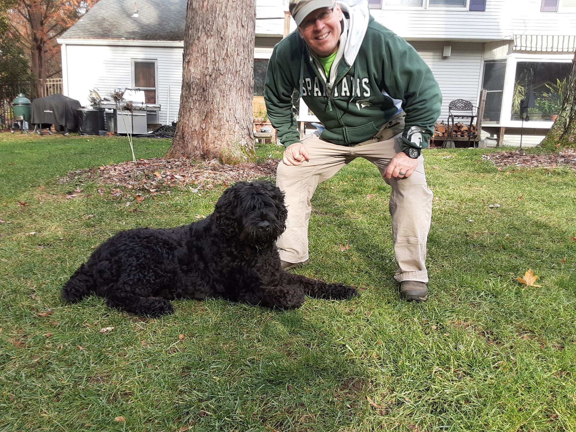 Black dog lying on grass beside a crouching person in a green sweatshirt in a yard