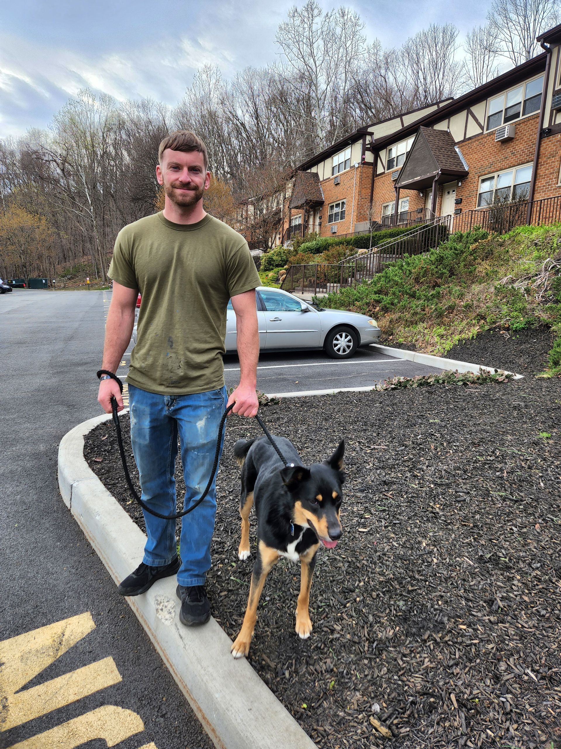 Man standing with a black-and-tan dog on a curb in a parking lot near houses and a parked car