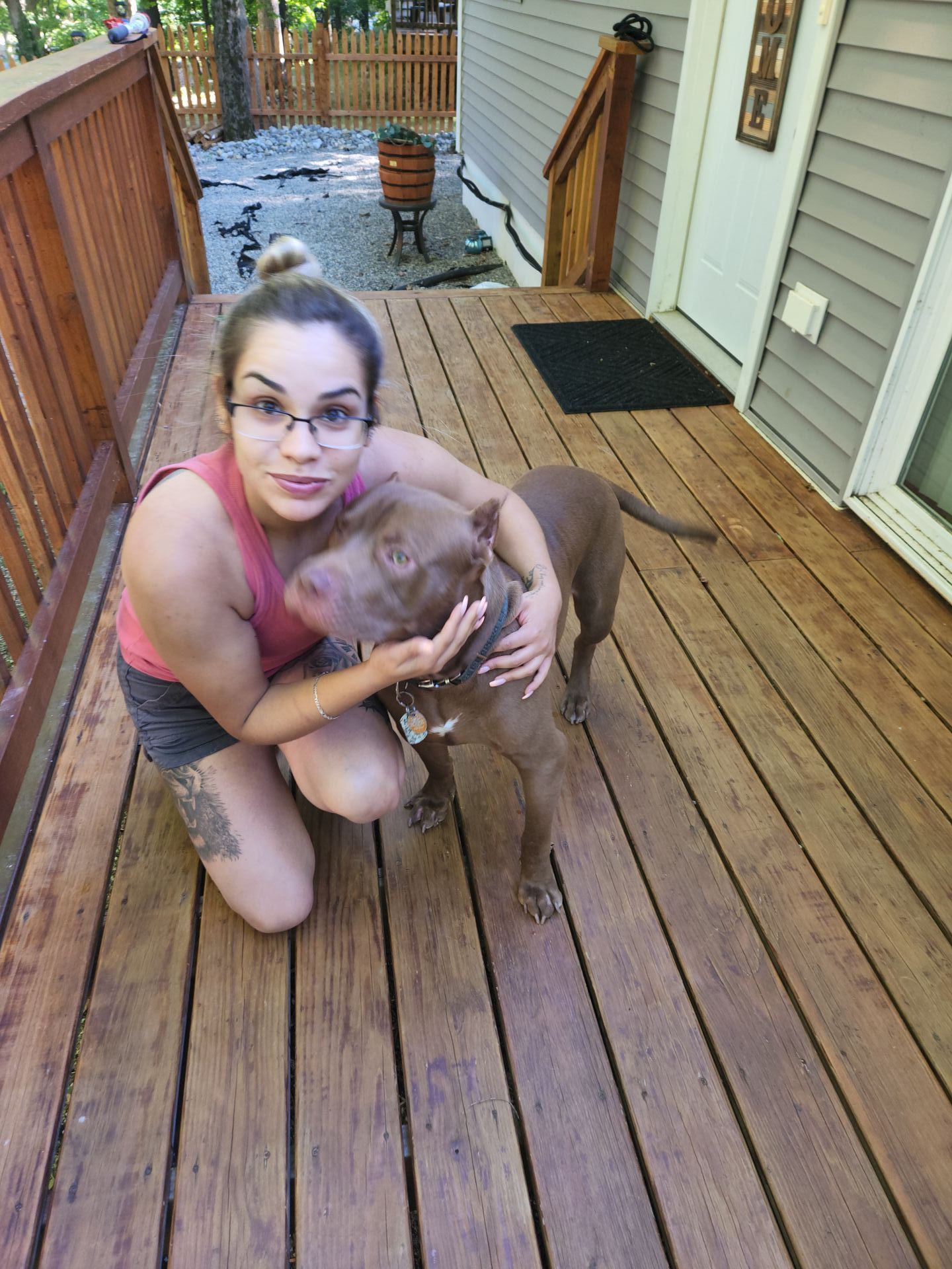 Woman kneeling on a wooden deck, hugging a brown dog outdoors near a house.