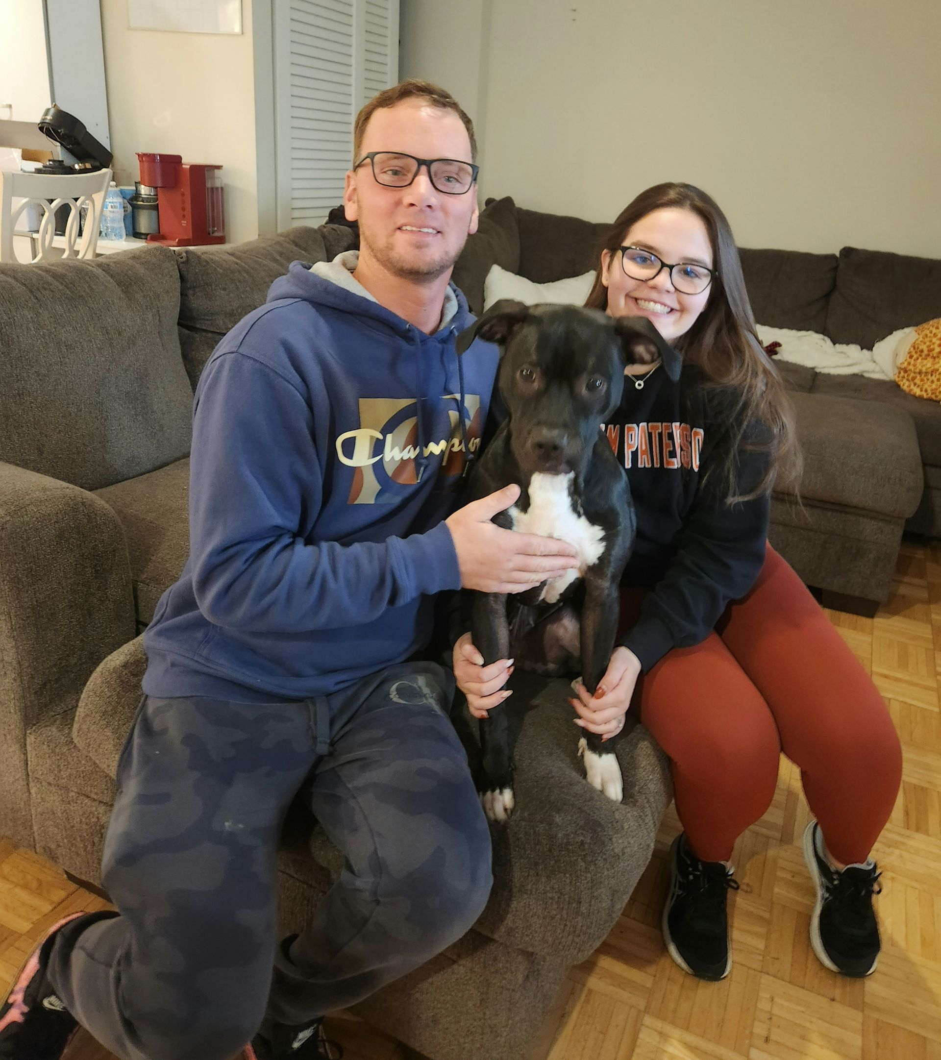 Two people sitting on a couch holding a black dog in a living room