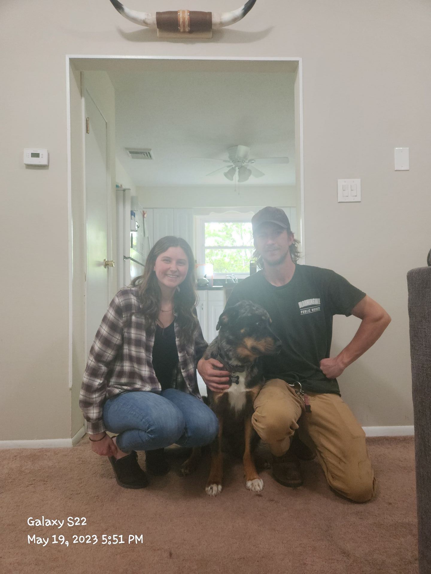 Two people kneeling with a dog in a doorway inside a carpeted living room