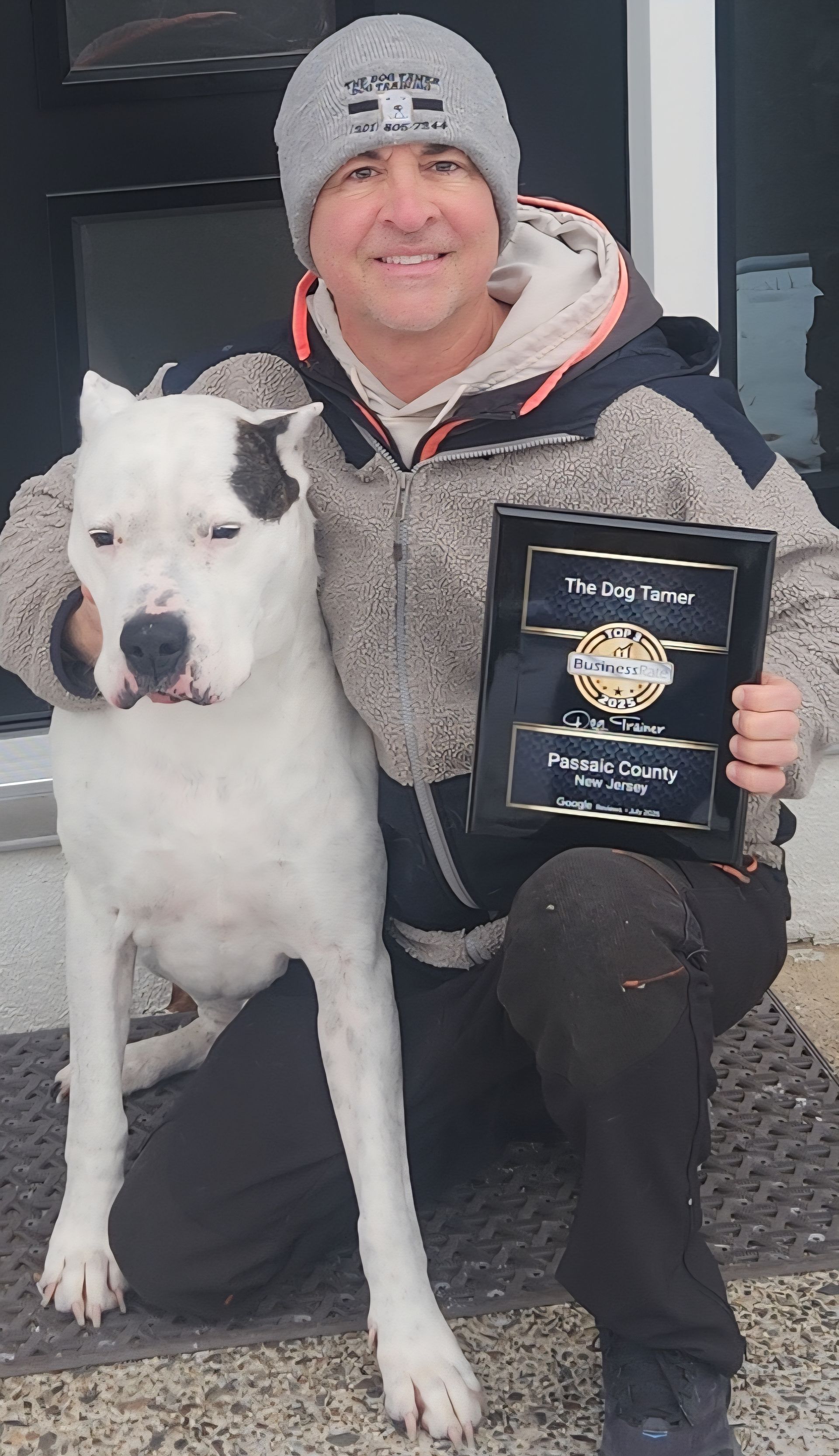 Person kneeling with a white dog, holding a framed award plaque indoors.