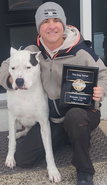 Person kneeling with a white dog, holding a framed award plaque indoors.