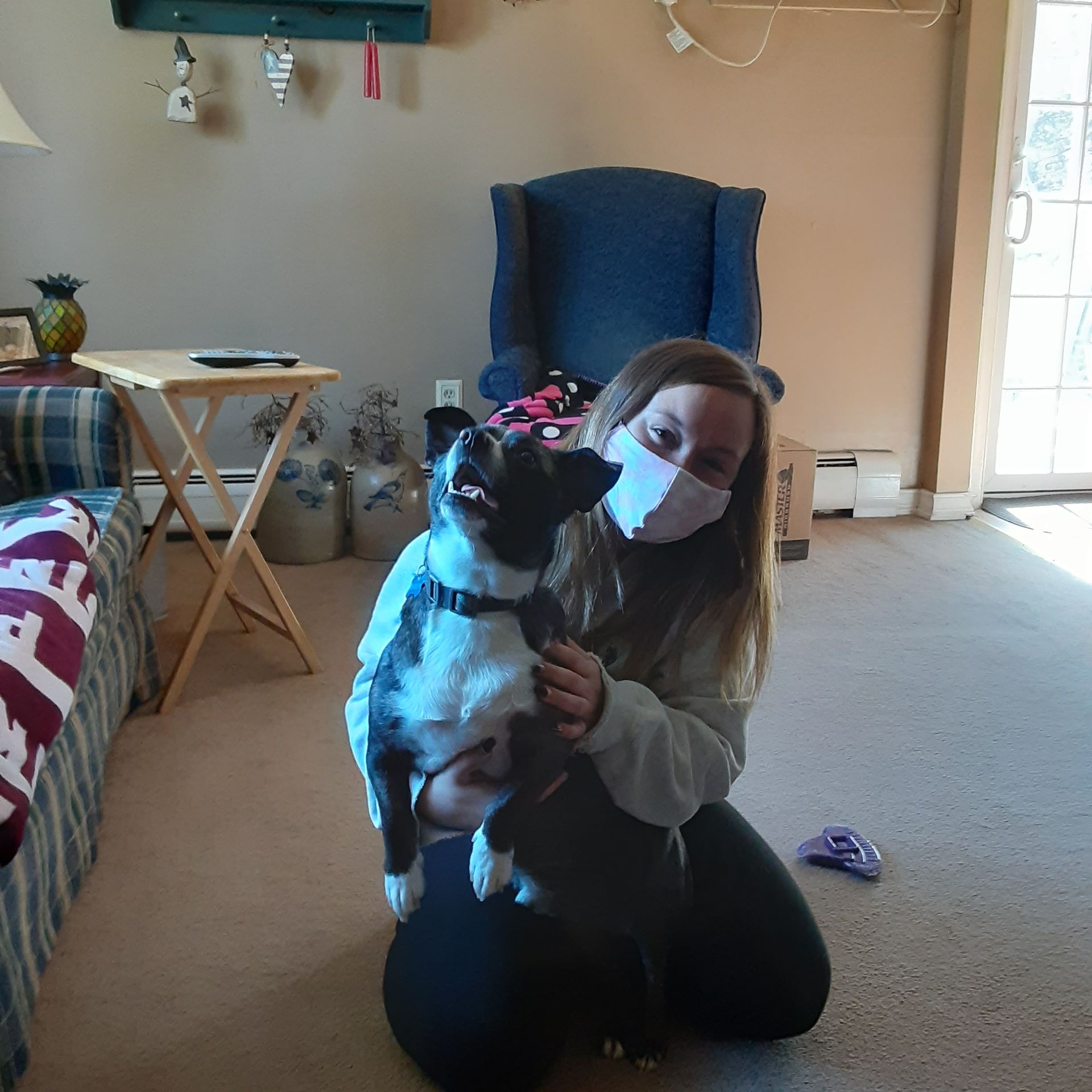 Woman kneeling indoors and holding a black-and-white dog, both wearing masks.