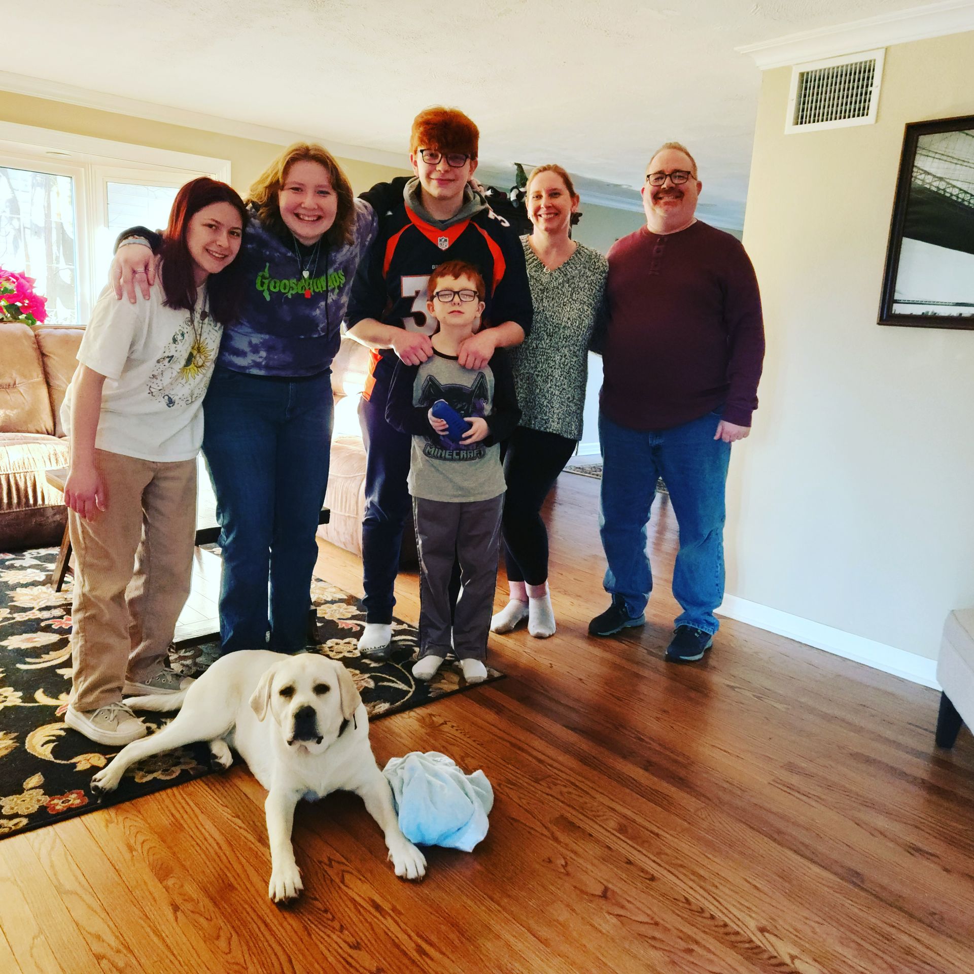 Group of six people posing indoors with a white dog lying on the floor in front.