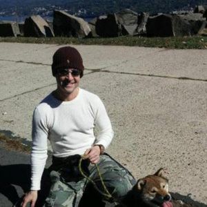 Smiling person in a beanie kneels on pavement beside a leashed dog outdoors.
