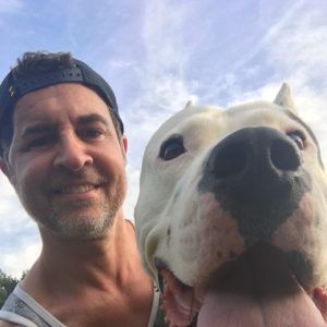 Smiling man in a backward cap with a close-up white dog against a blue sky