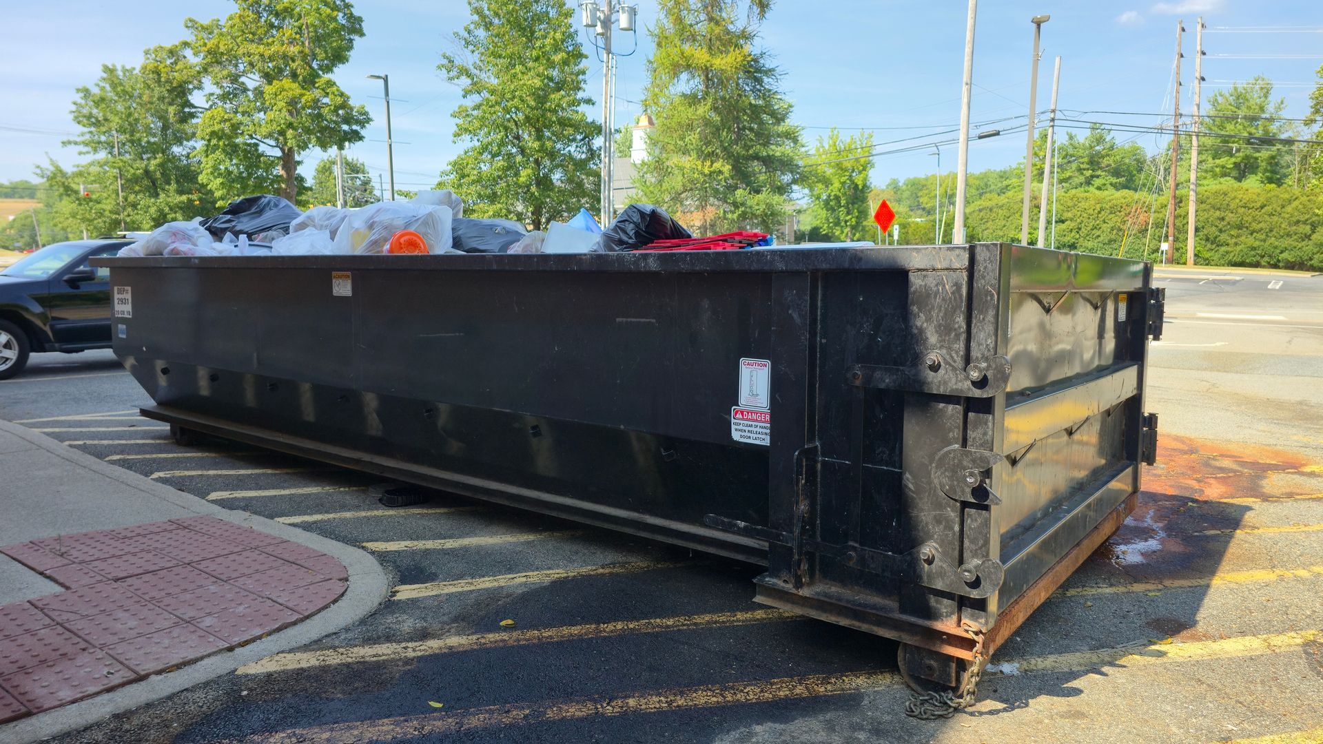 Green dumpster full of trash beside a yellow wall, with a traffic cone nearby.