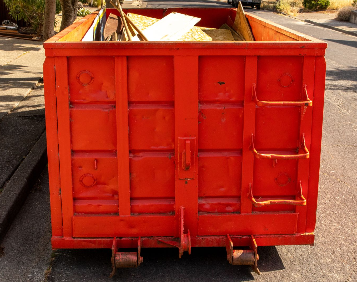 Orange construction truck behind blue dumpsters on a street, with debris and an excavator arm visible.
