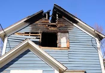 Blue house with severe fire damage around the roof and an open, blackened attic window