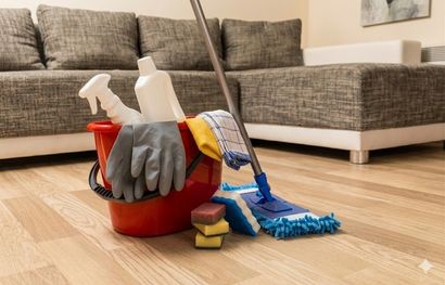 Cleaning supplies on a hardwood floor with a mop, bucket, spray bottles, and a couch in the background