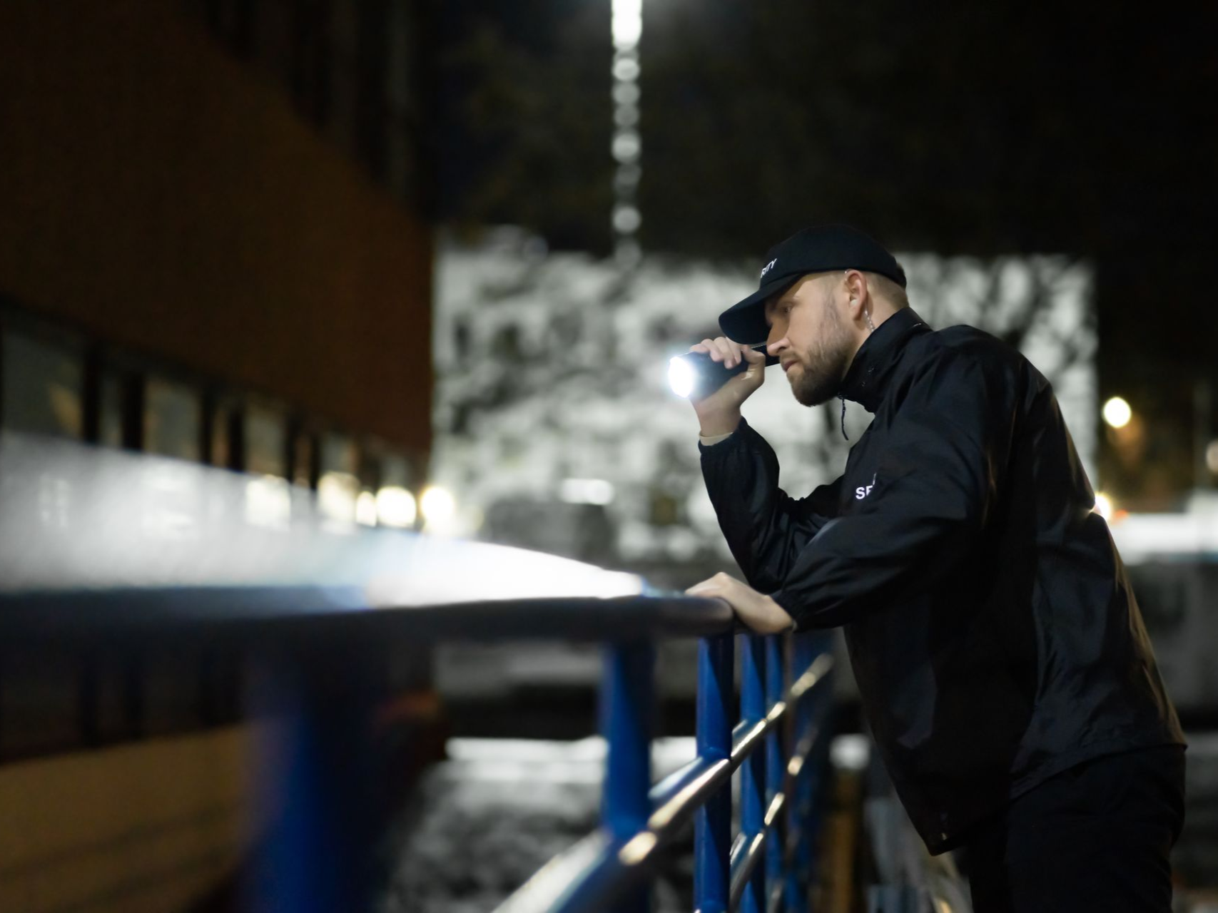 Person leaning on a blue railing at night, drinking from a cup in a city setting.