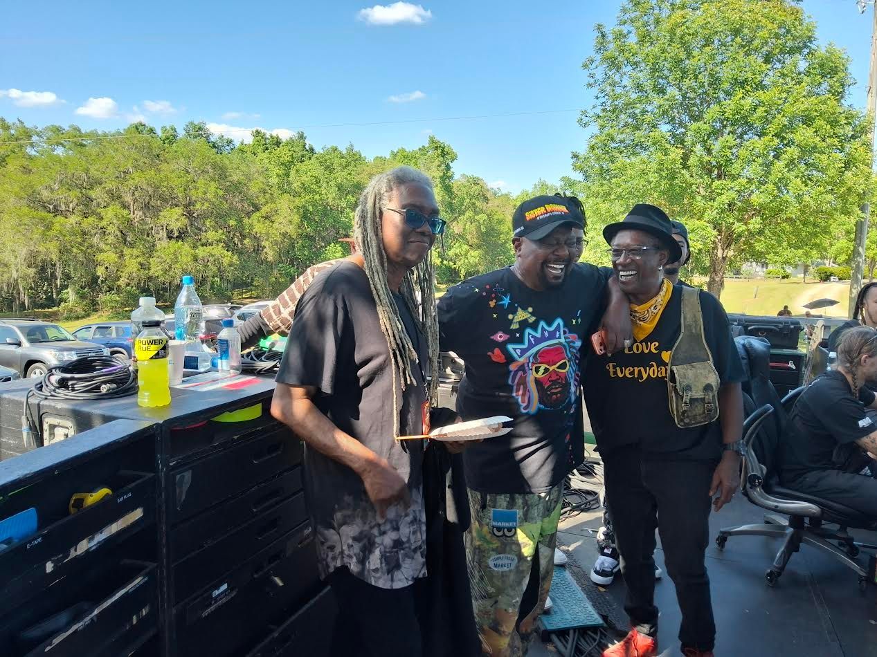 Three people posing beside a picnic table in a sunny park with trees and drinks nearby