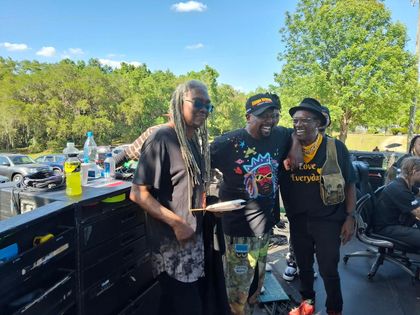 Three people posing beside a grill at a sunny outdoor cookout, with trees and tables in the background