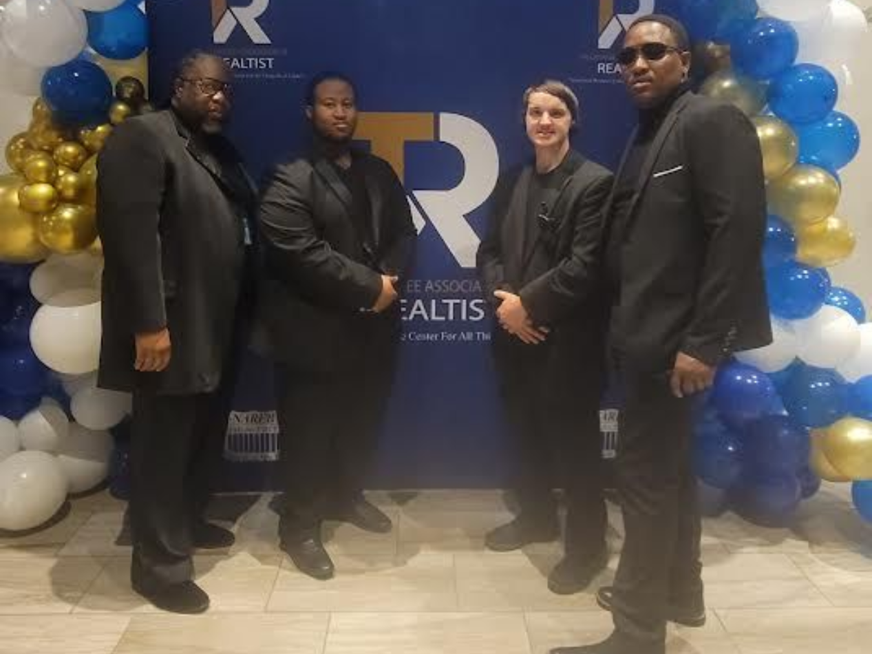 Four people in suits pose in front of a blue-and-gold balloon backdrop at an event.