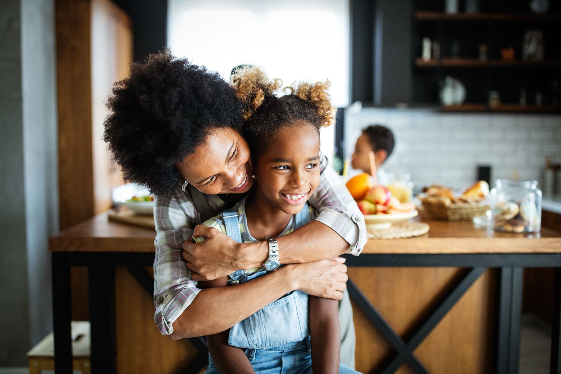 A woman is giving a little girl a piggyback ride in the kitchen.
