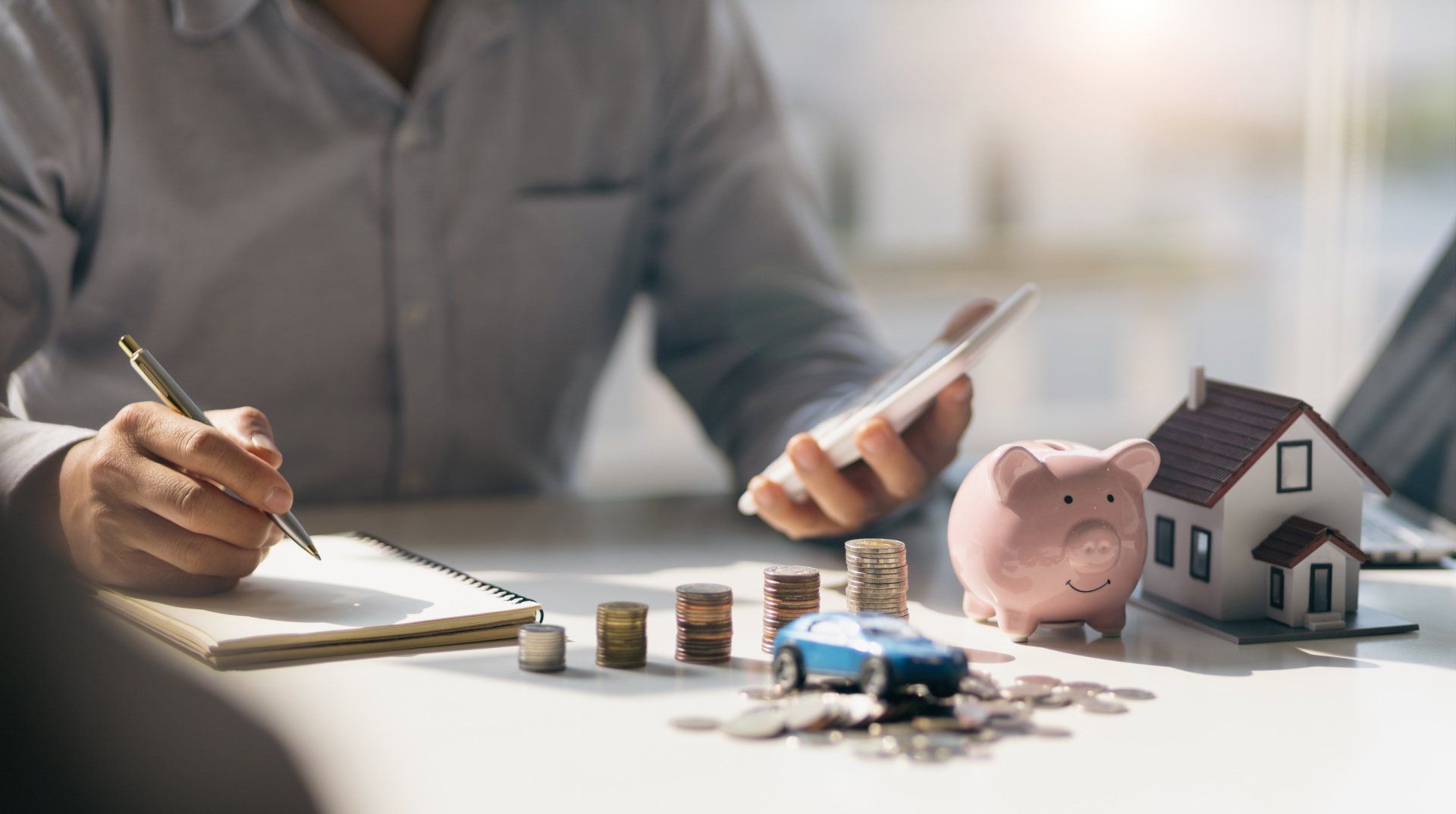 A man is sitting at a table with a piggy bank , coins , a car , and a house.