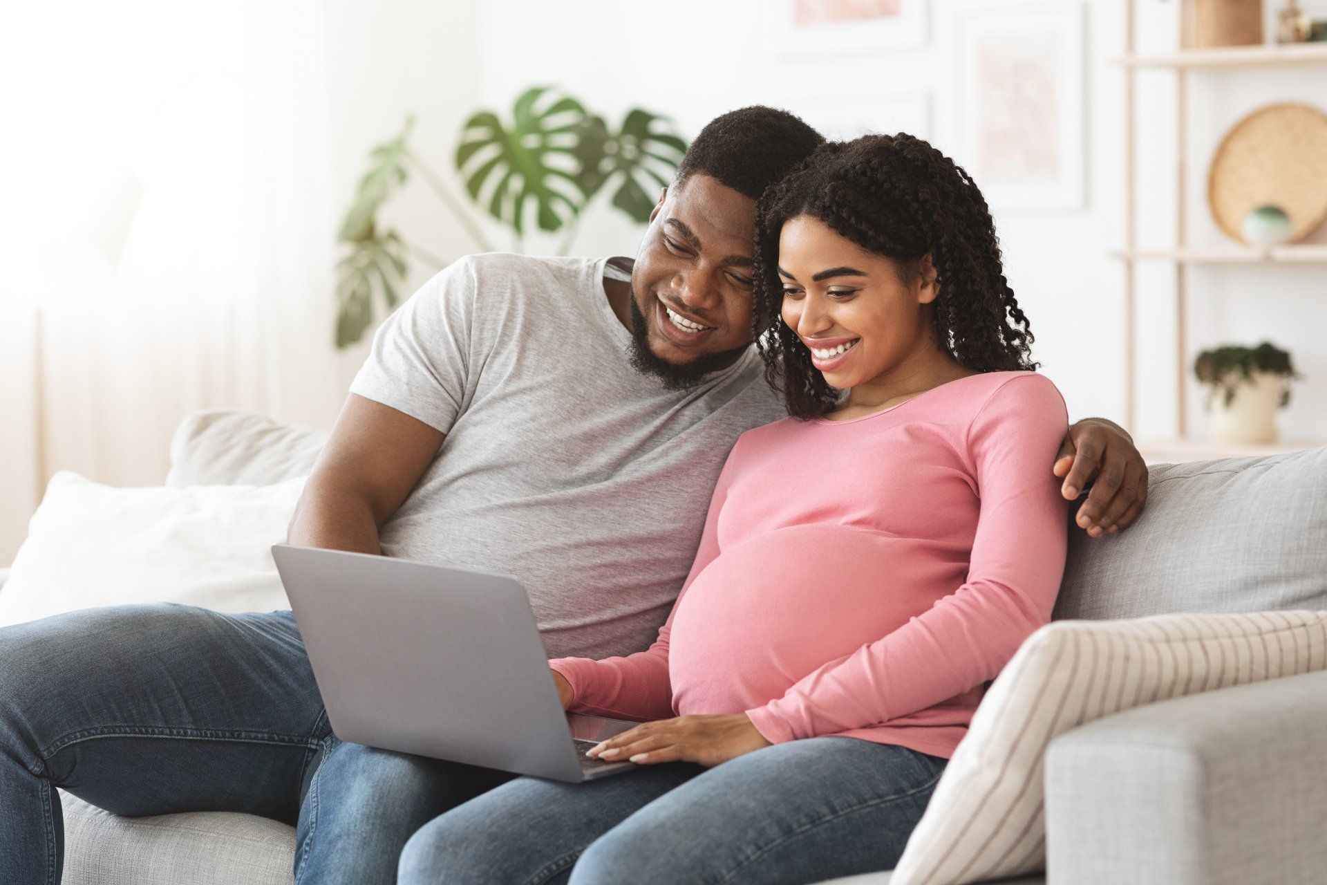 A man and a pregnant woman are sitting on a couch looking at a laptop.