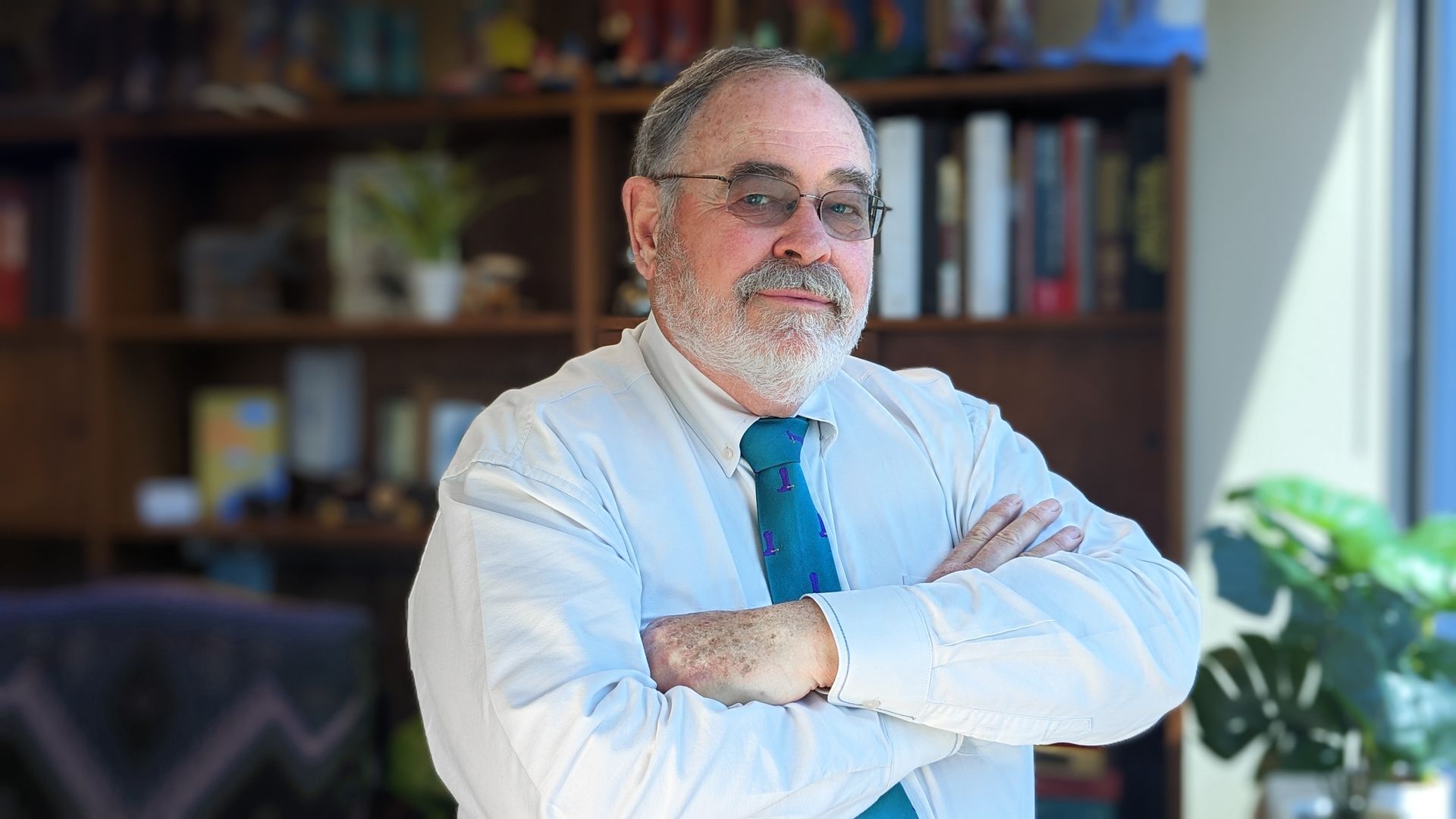 A man with a beard and glasses is standing with his arms crossed in front of a bookshelf.