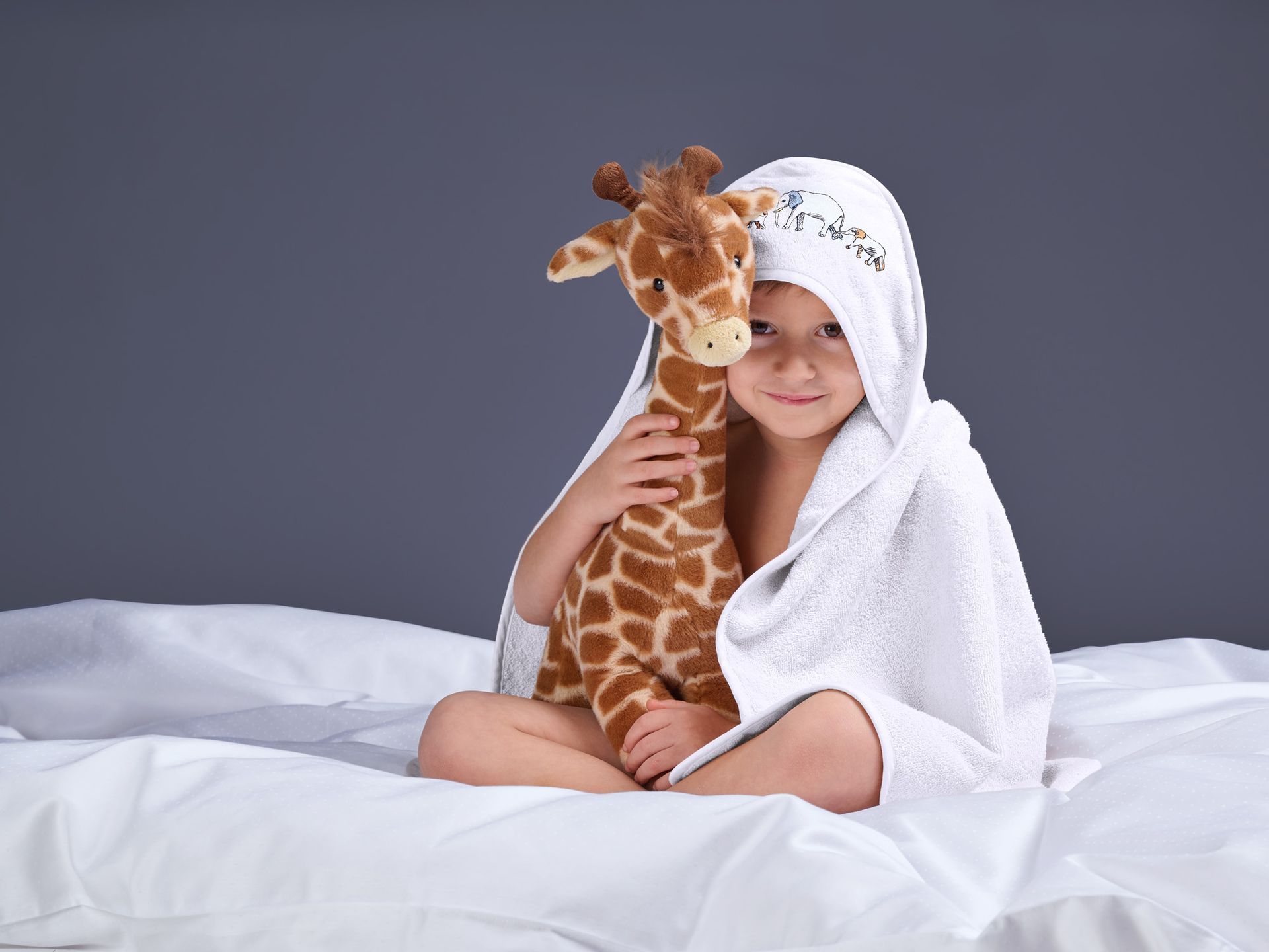 Child in a white bathrobe sitting on a bed, hugging a stuffed giraffe and smiling.
