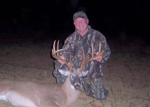 Hunter kneels beside a large buck at night, wearing camouflage in a dark field.