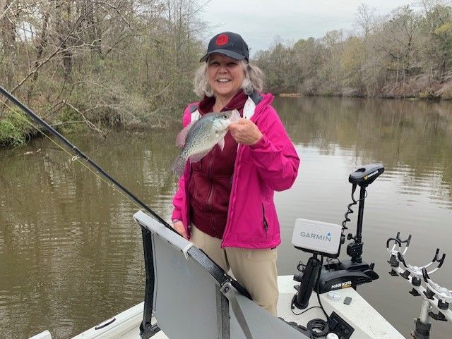 Person holding a fish on a boat beside a calm river, wearing a pink jacket and black cap