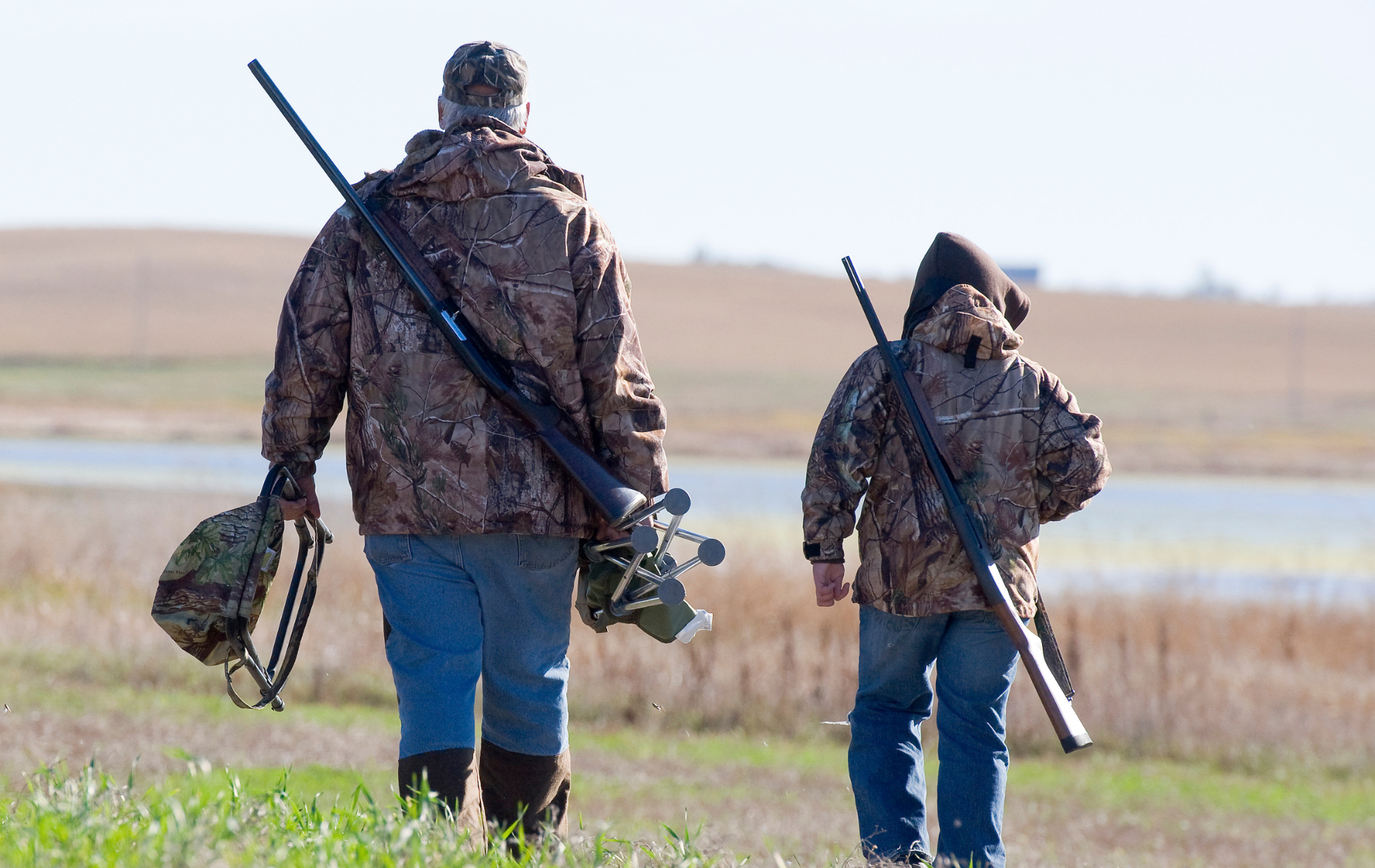 Two hunters in camouflage walking across a field with shotguns and gear on their backs