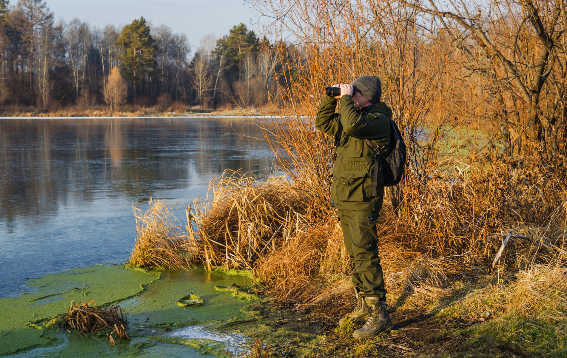 Person photographing a misty riverbank with autumn trees and green algae on the water