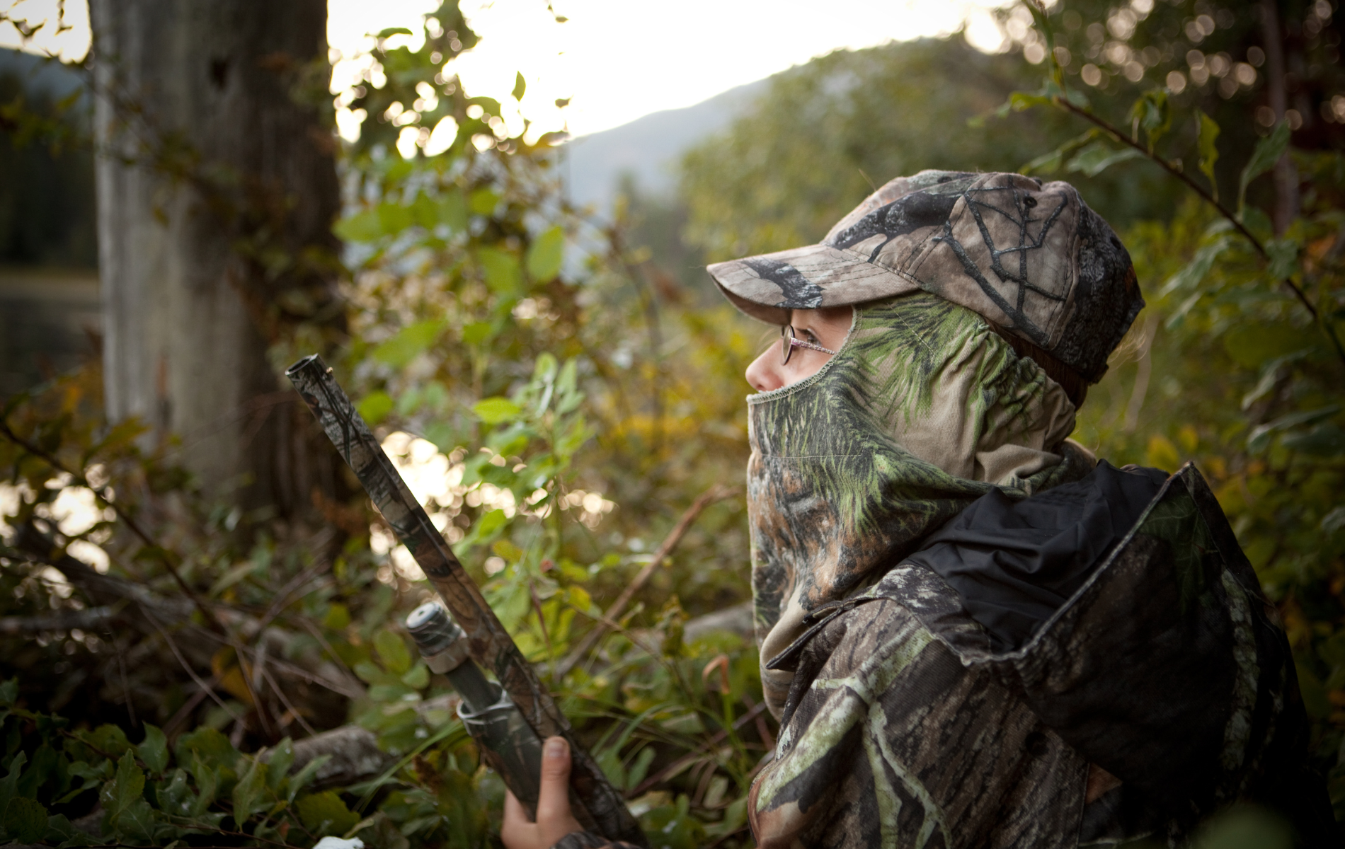 Camouflaged hunter aiming a rifle in a wooded area from behind a tree