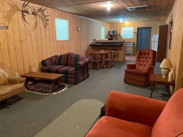 Basement living room with wood-paneled walls, couch, armchairs, bar stools, and carpet.