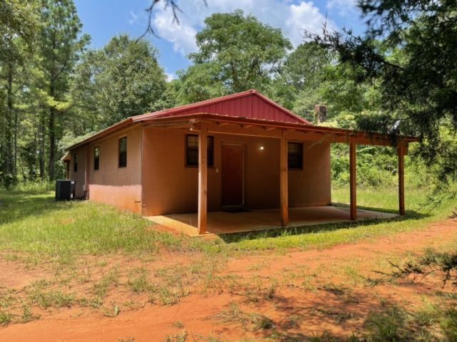 Small tan house with red roof and porch, surrounded by trees and grassy yard.