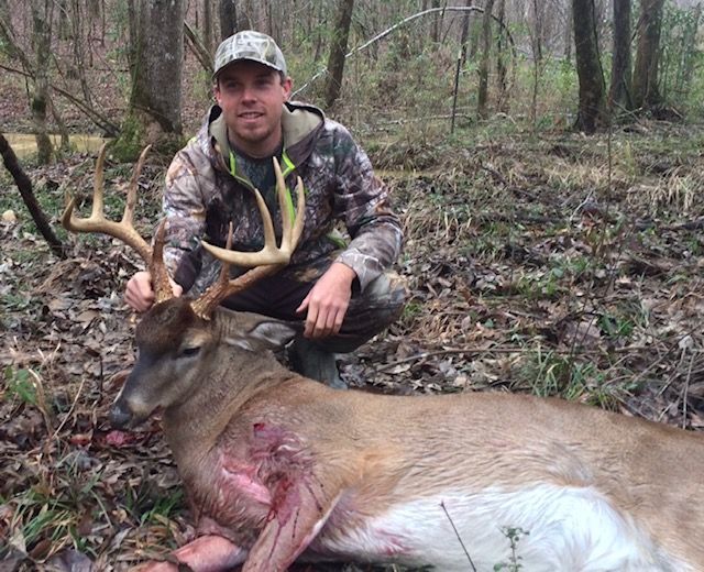 Hunter kneeling beside a large deer in a wooded area, with the deer’s antlers visible.