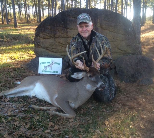 Hunter posing with a deer in a wooded area beside a large tree stump at sunset.