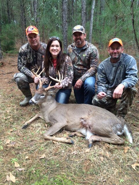 Four hunters kneel behind a large deer in a wooded clearing, holding the antlers.