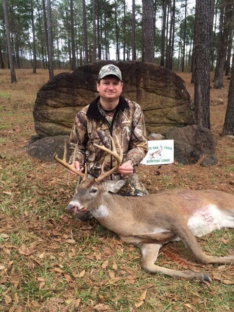 Hunter kneels beside a large deer in a pine forest, holding its antlers.