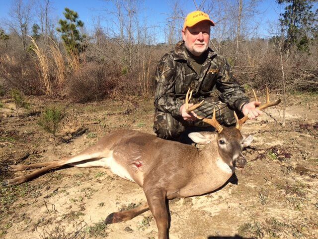Hunter kneeling behind a deer in a dry field, holding the antlers, wearing camouflage and a yellow cap