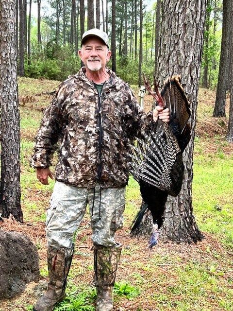 Man in camouflage holding a large turkey by a pine tree in the woods