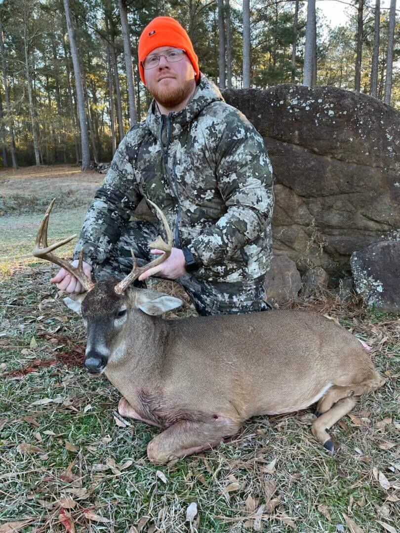 Hunter in orange beanie kneels beside a deer in a wooded area, holding its antlers.