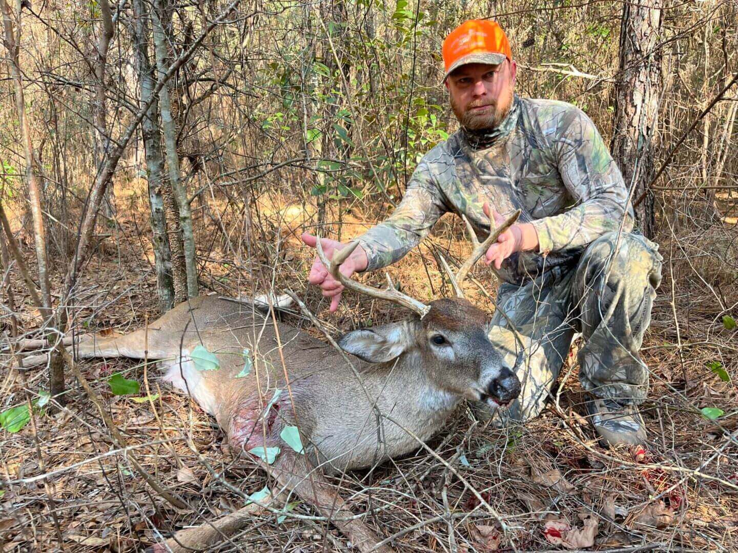 Hunter kneels beside a deer in a wooded area, wearing camouflage and an orange cap.
