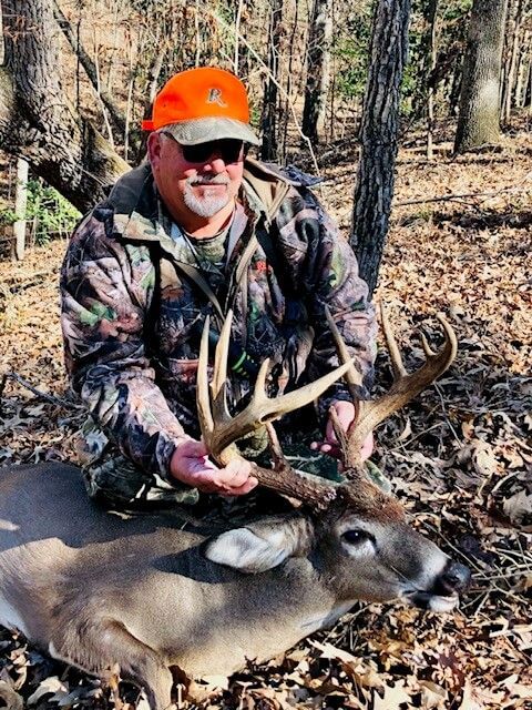 Hunter in orange cap kneeling beside a deer in a wooded area, holding large antlers.