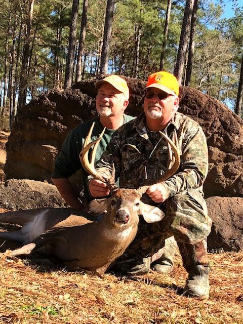 Two hunters kneel beside a large deer in a wooded area, wearing orange caps and camouflage.