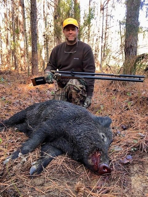 Hunter kneels behind a large wild boar in a wooded area, holding a scoped rifle.