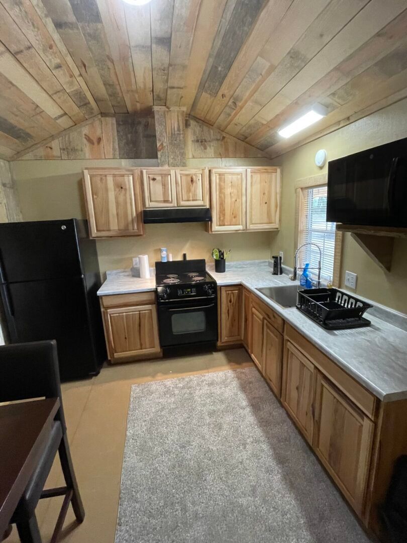 Small kitchen with wood cabinets, black appliances, gray countertop, and a rug under a wood plank ceiling.