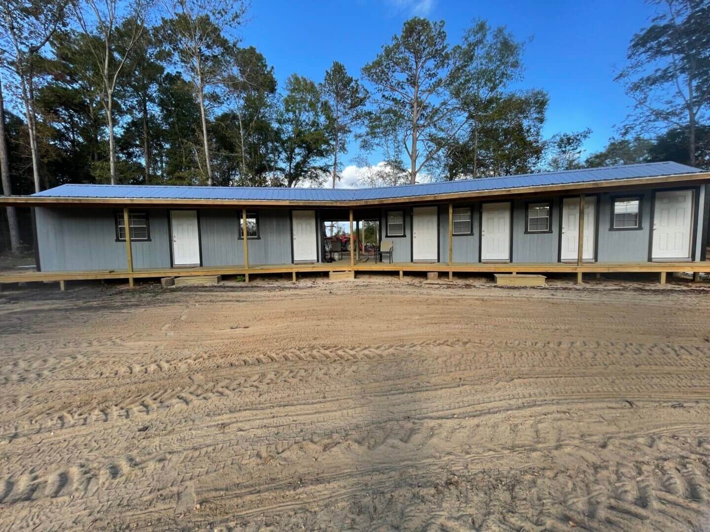 Single-story blue building with a long porch and trees behind it, on a dirt lot.