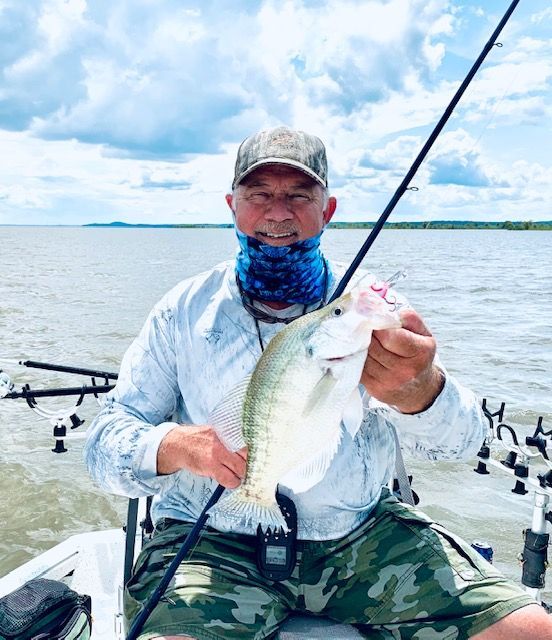 Angler on a boat holding a large fish, with rods and calm water under a cloudy sky.