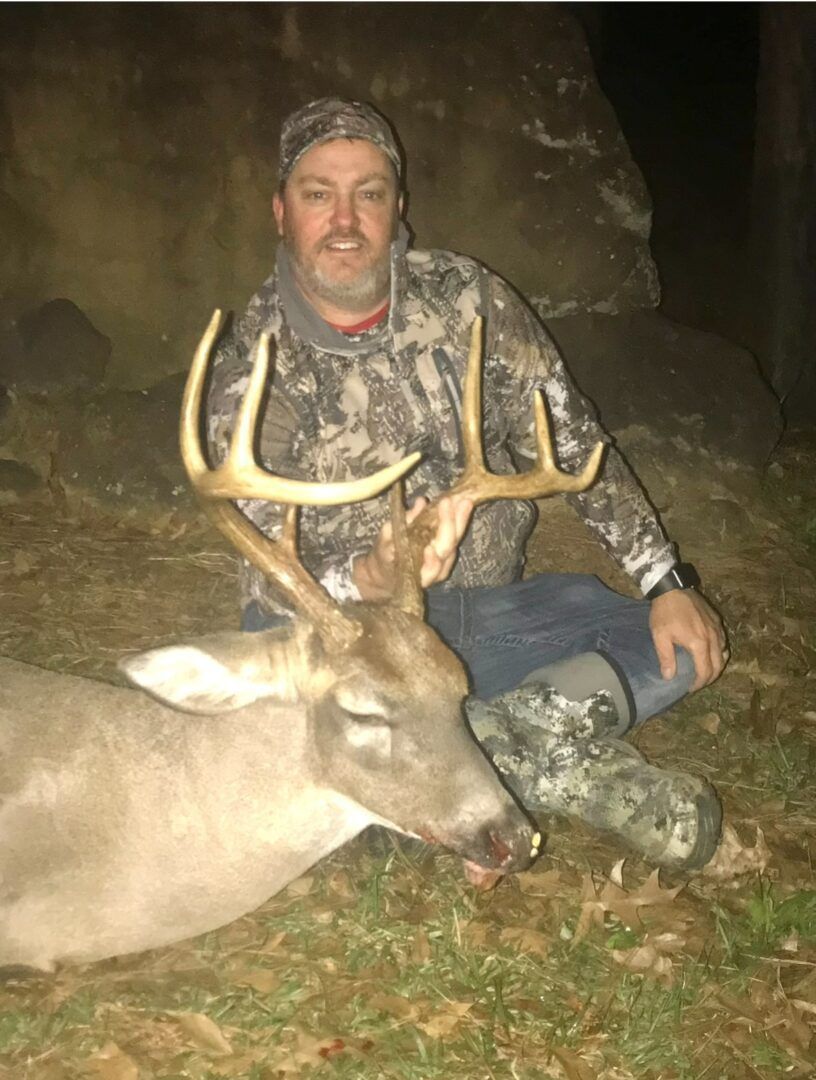 Hunter kneels beside a large deer at night, wearing camouflage in a wooded area.