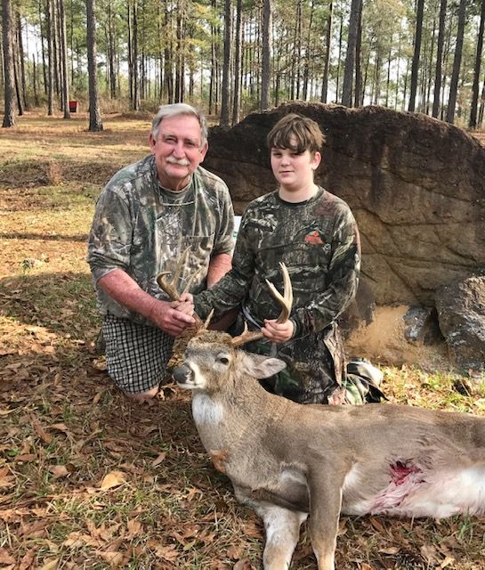 Two people in camouflage pose with a deer in a wooded area after a hunt.