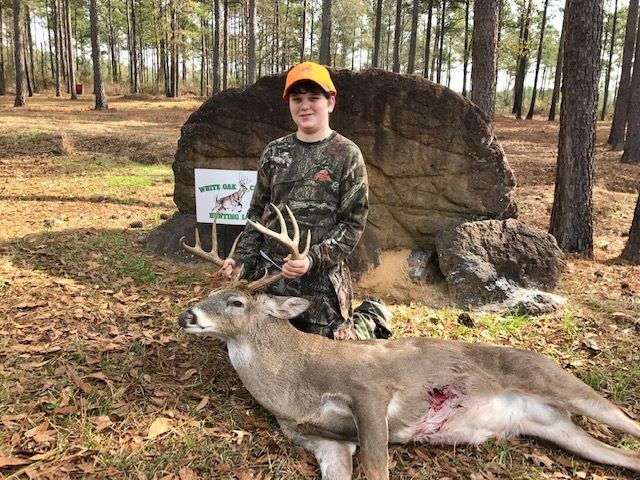 Youth in camo poses with a deer beside a rock marker in a pine woods clearing.